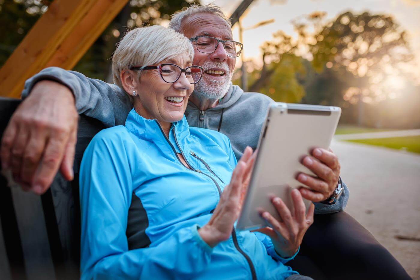 Lächelndes älteres aktives Paar, das auf einer Bank im Park sitzt und auf einen Tablet-Computer schaut. Nutzung moderner Technologie durch ältere Menschen